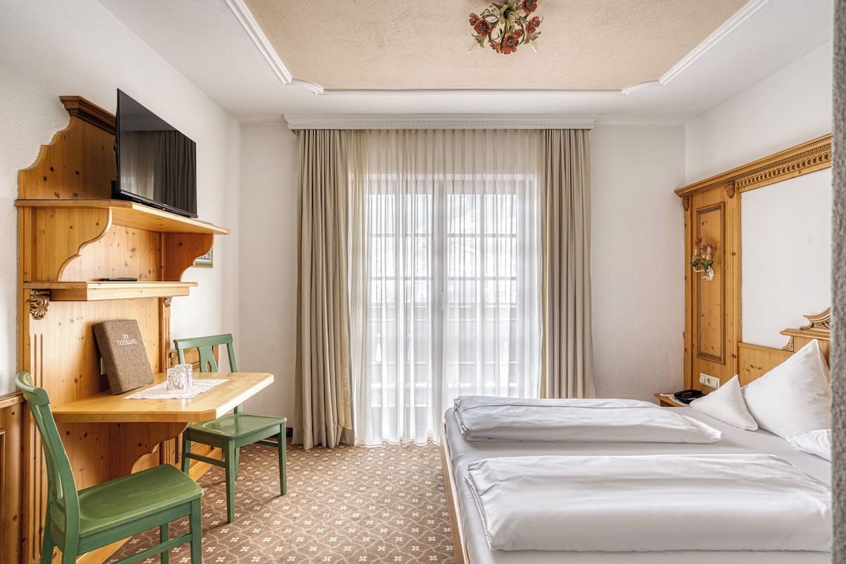A hotel room with a bed, a desk with a chair, a television, and a large window with curtains.