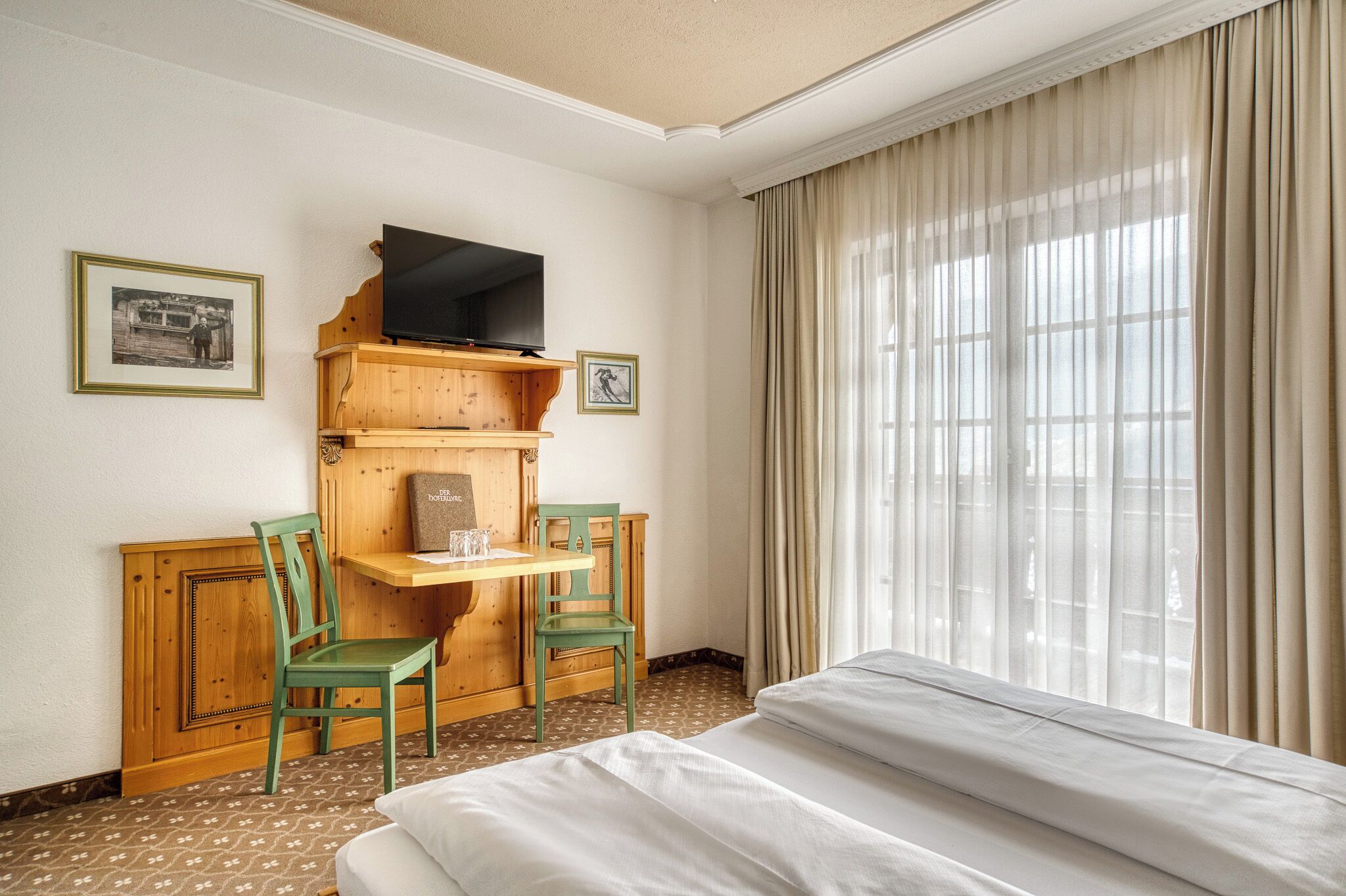 A hotel room with a bed, a wooden desk with a television, two chairs, and a window with curtains.