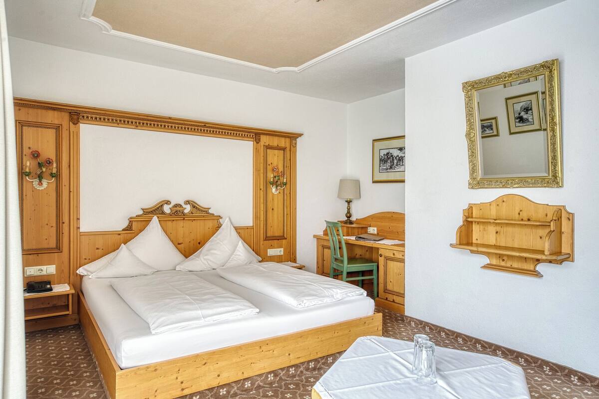 A hotel room with a wooden bed, a desk with a chair, a mirror, and a small shelf.