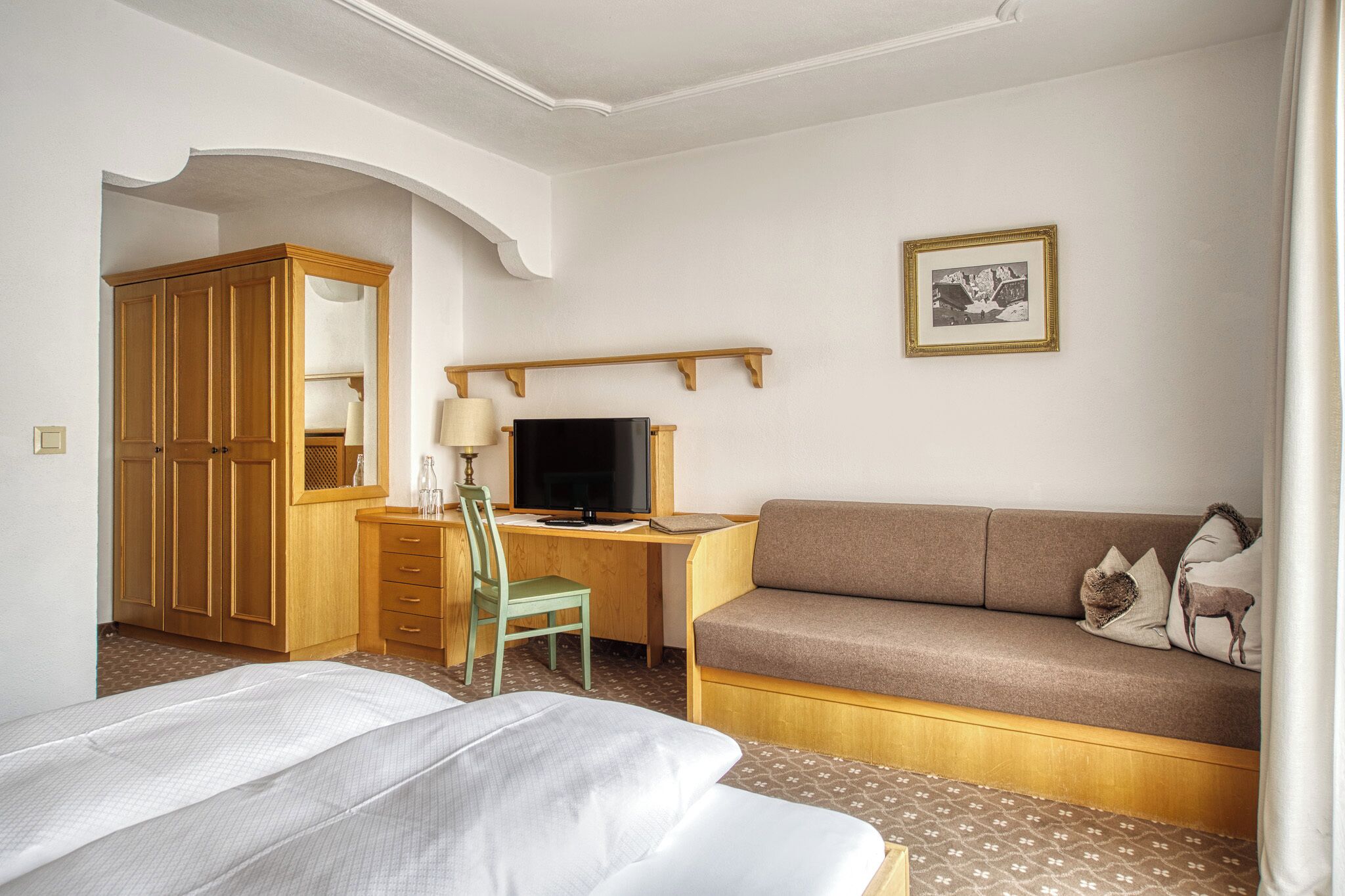 Quarto de hotel com uma cama, uma escrivaninha com computador, um sofá, uma poltrona e um guarda-roupa.