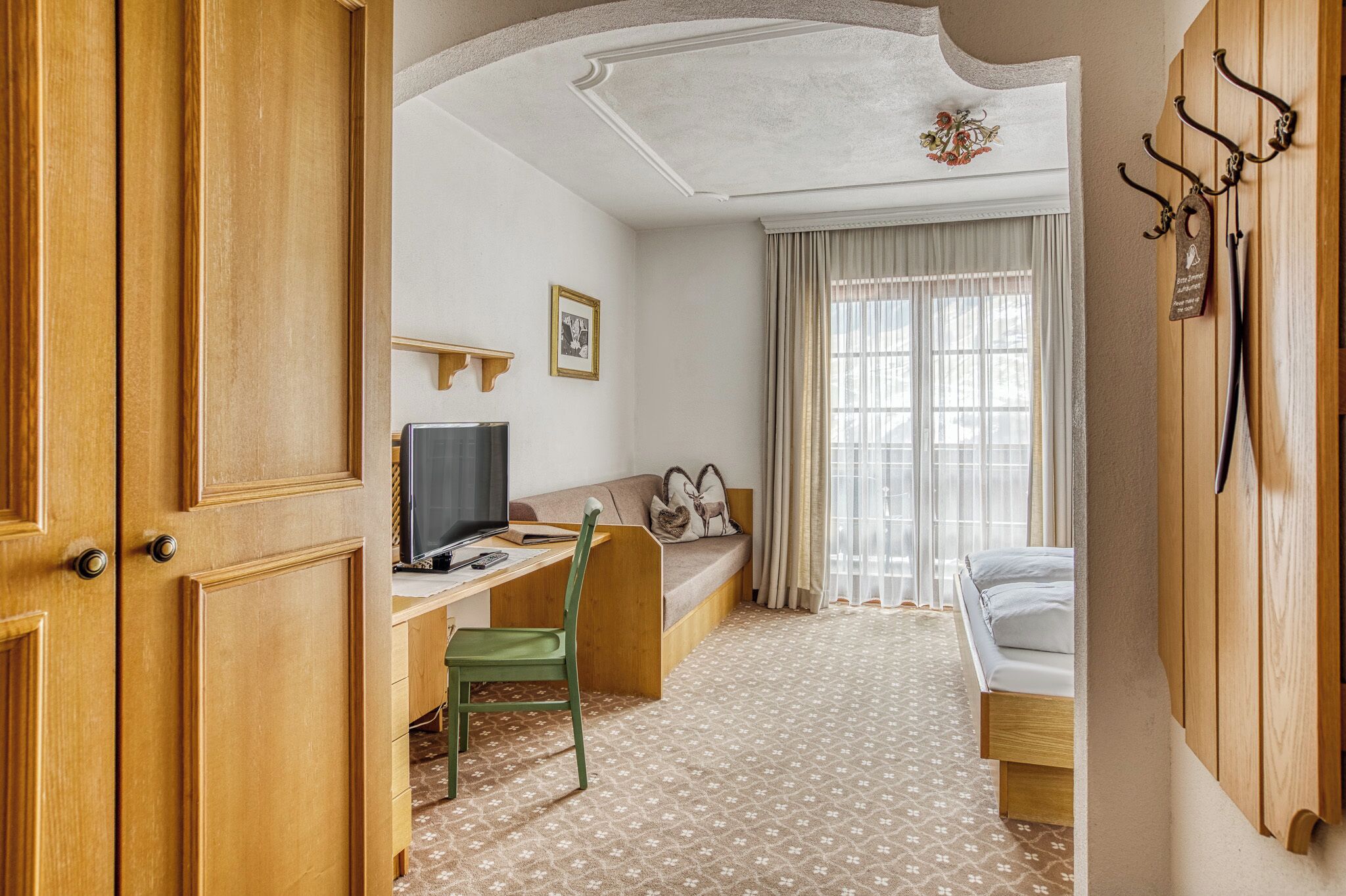 A hotel room with a wooden desk, a green chair, a bed, a TV, and a window with curtains.