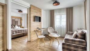 A modern hotel room with a wooden floor, a bed, a sofa, a chair, a small table, a TV mounted on the wall, and a large window with curtains. - Der Hoferwirt (Neustift Im Stubaital)