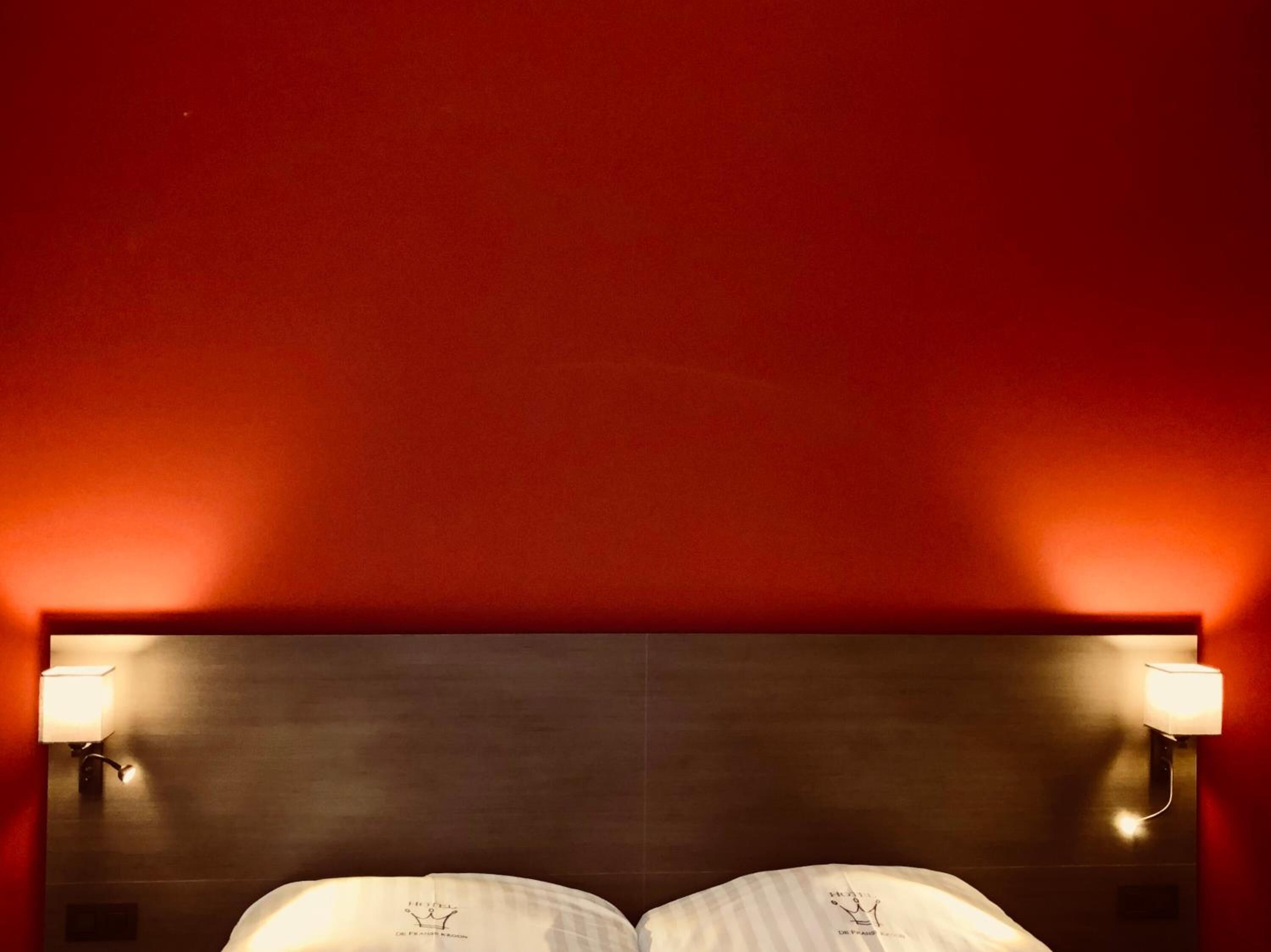 Habitación de hotel con cabecera oscura, dos lámparas de noche y un logotipo visible en la cabecera.