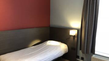 Un lit simple dans une chambre dont un mur est peint en rouge et en gris, avec une lampe de chevet et une fenêtre avec des rideaux.