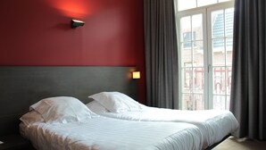 A hotel room with two beds, a red wall, a window with curtains, and a view of a building outside. - Hotel De Franse Kroon (Diest)