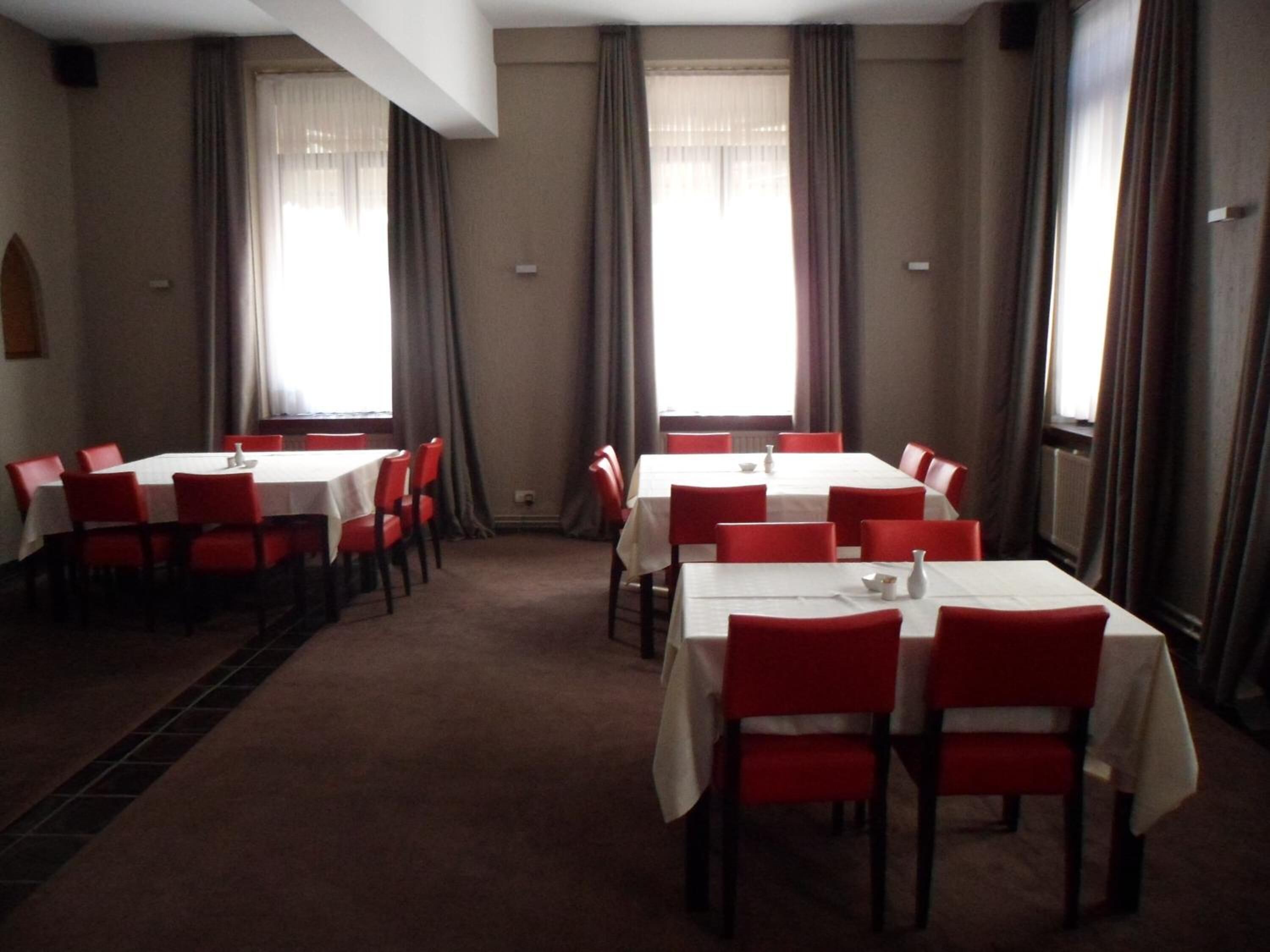 Une salle de conférence avec des tables blanches et des chaises rouges, de grandes fenêtres avec des rideaux et un sol recouvert de moquette.