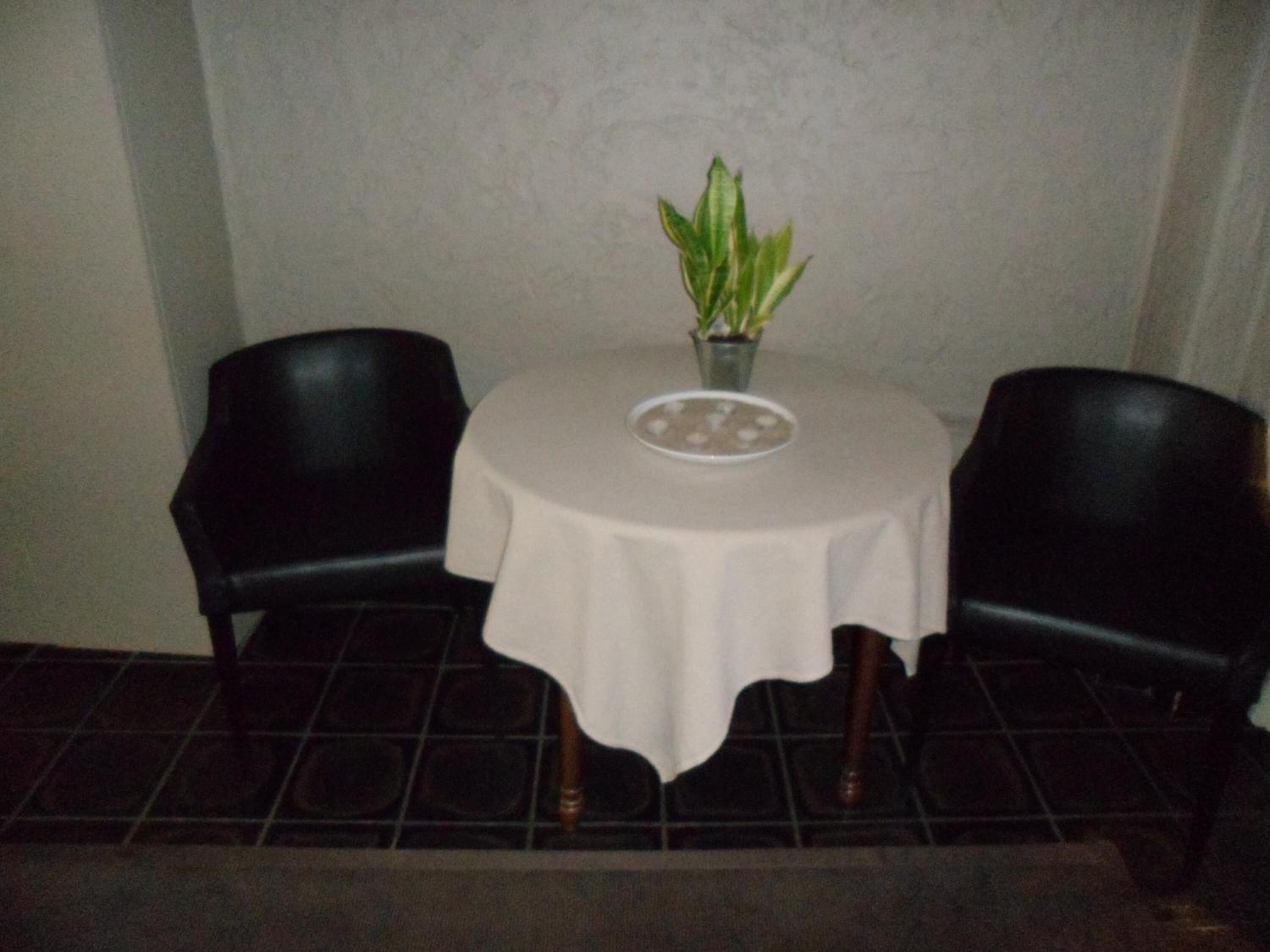 Ein kleiner Essbereich mit einem runden Tisch, der mit einer weißen Tischdecke bedeckt ist, zwei schwarzen Stühlen und einem Blumentopf auf dem Tisch.