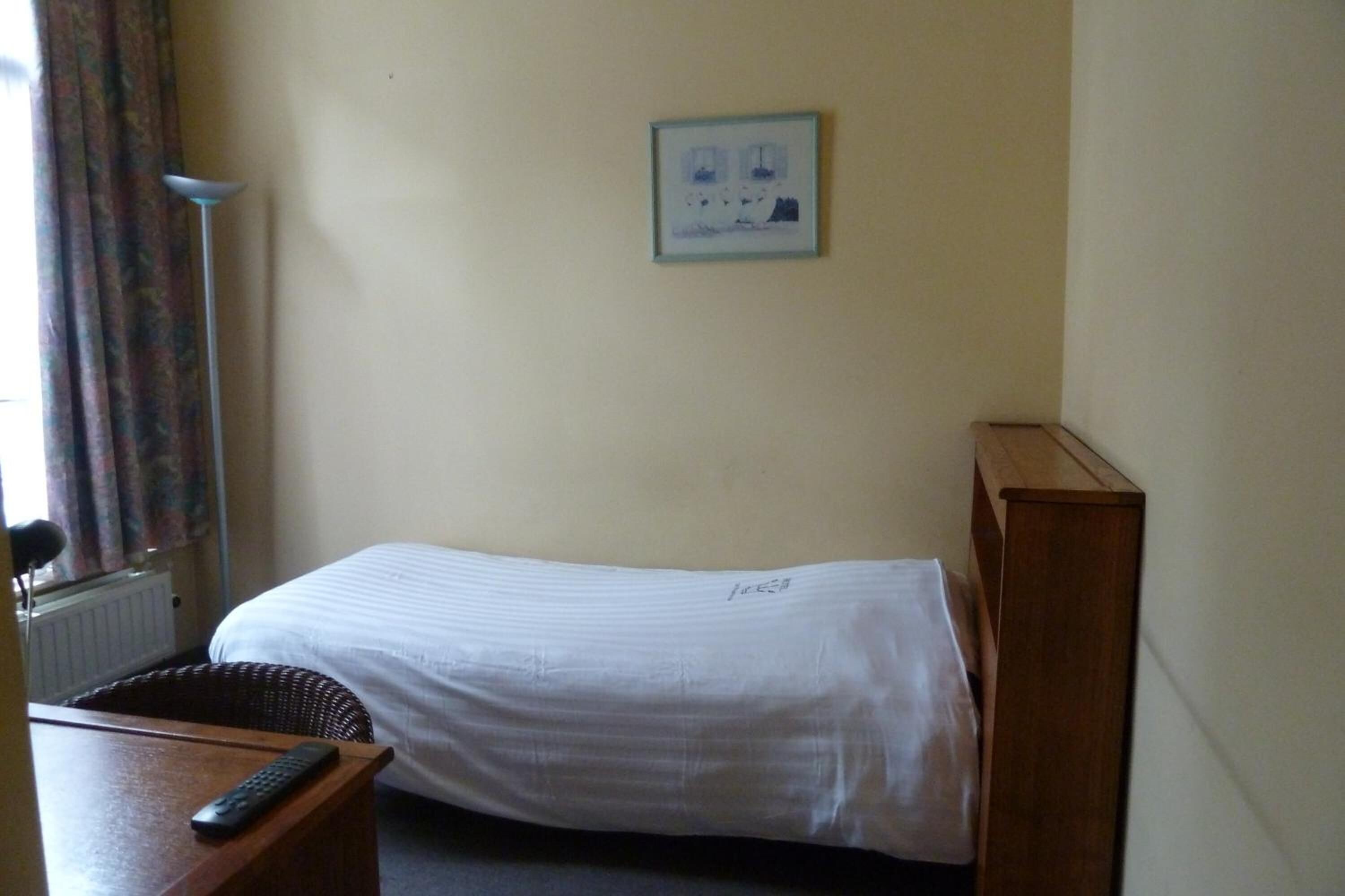 Una cama individual con colchón y cabecera blancos, una mesita de noche con control remoto, un cuadro en la pared y una ventana con cortinas.