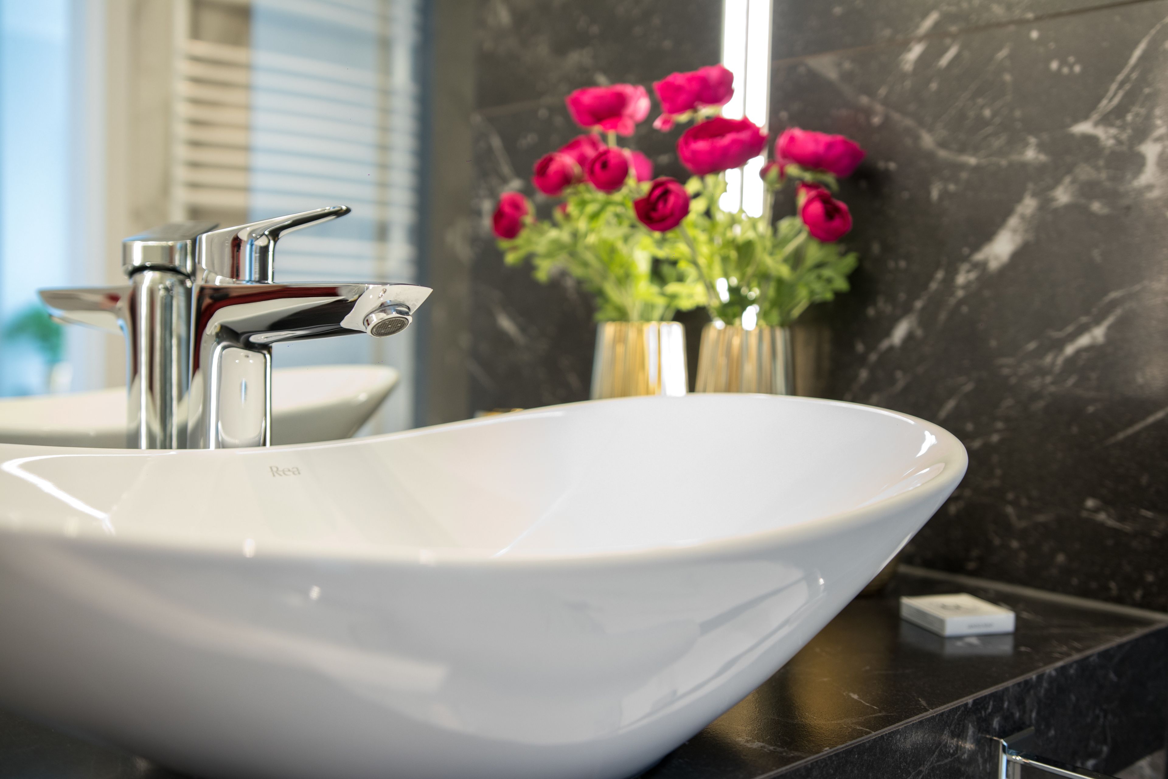 Un baño con un lavabo blanco tipo recipiente, grifo cromado y un florero con flores rosadas sobre la encimera.