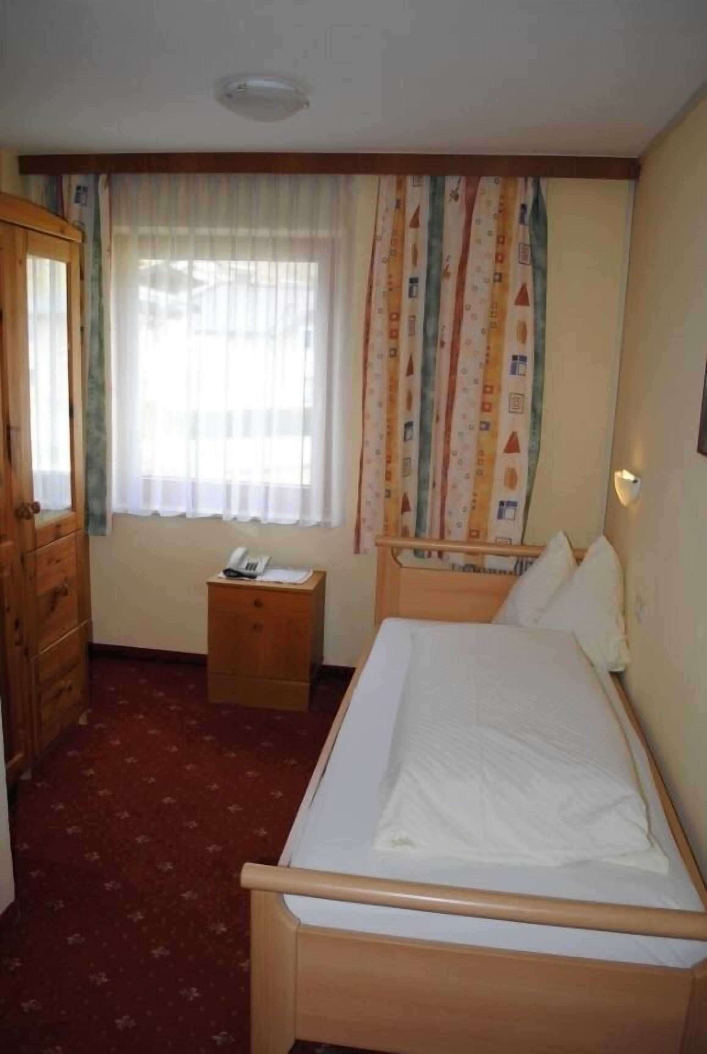 A room with a single bed, a wooden wardrobe, a bedside table with a lamp, and a window with curtains.