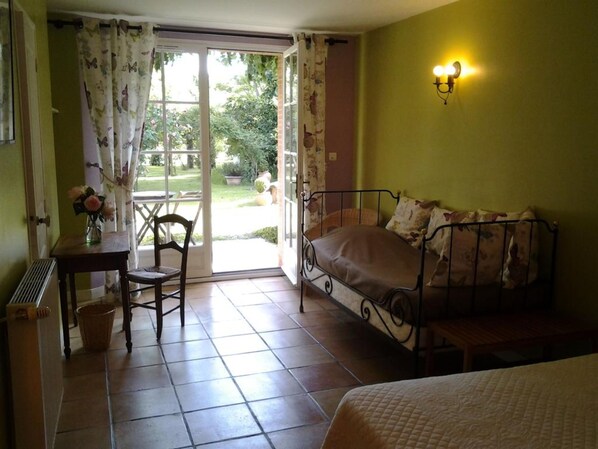 Un dormitorio con cama, una silla, una mesita con un florero y una ventana con cortinas.