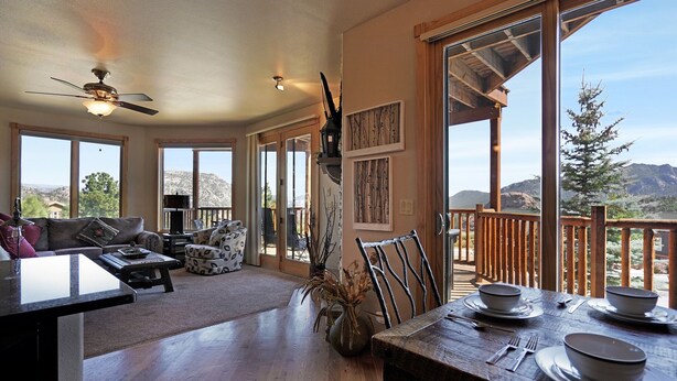 Un vaste espace de vie comprenant un coin repas avec une table pour quatre personnes, un ventilateur de plafond et de grandes fenêtres offrant une vue sur les montagnes.