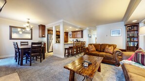 A spacious living room with a wooden coffee table, a brown leather sofa, and a dining area with wooden chairs and a kitchen in the background. - Horizons 4 165 Updated Condo, Walk to Town, On Shuttle Route to Lodges by RedAwning (Mammoth Lakes)