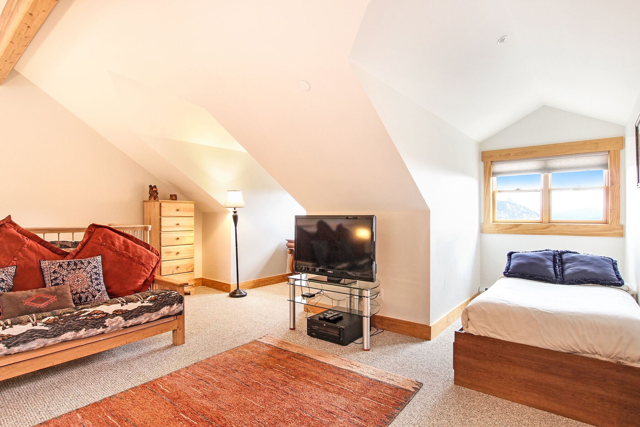 Ein gemütliches Schlafzimmer im Loft-Stil mit schrägem Dach, einem Bett, einer Couch, einem Fernseher auf einem Glastisch und einem Fenster mit Bergblick.