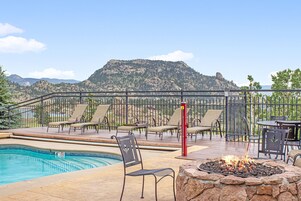 Zona de piscina con sillones de descanso y un hogar para fuego, rodeada por una terraza y vista a la montaña.