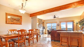 A dining area with wooden chairs and a table, a kitchen with a wooden bar, and a view of mountains through large windows.