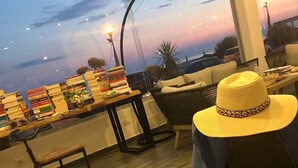 A cozy reading nook with a large window overlooking a cityscape, a stack of books, and a hat on a chair. - Drymades Resort (Himarë)