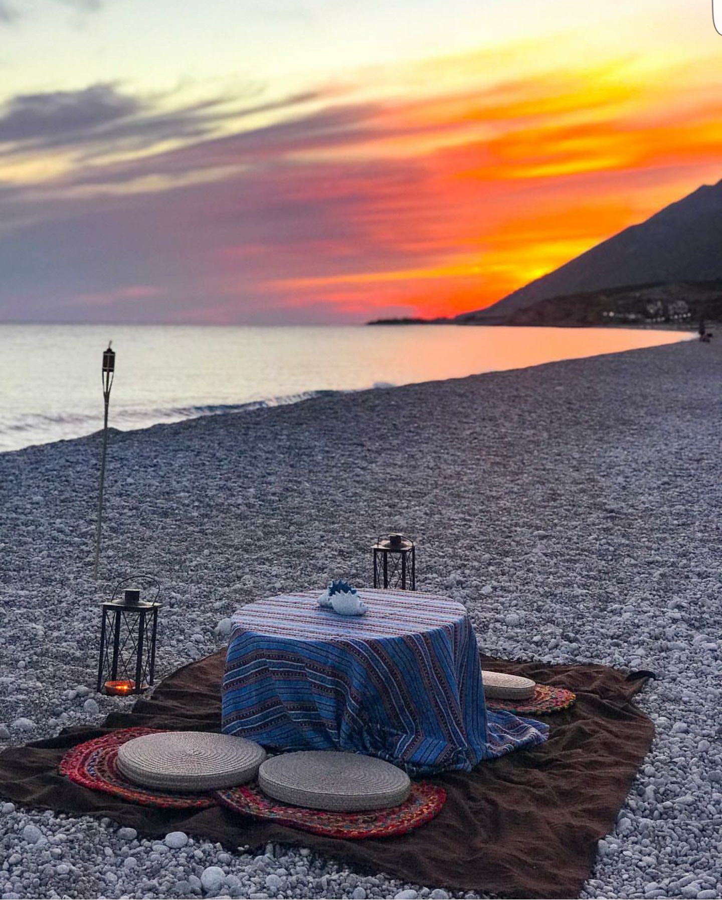A beach setting with a table set for a meal, two lanterns, and a blanket.