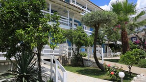 A hotel with a white and blue facade, a covered outdoor area, and a garden with various plants and a palm tree. - Drymades Resort (Himarë)