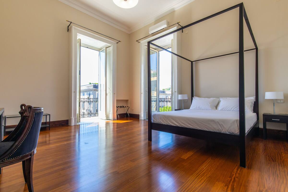 A spacious bedroom with a four-poster bed, a wooden chair, and a balcony view.