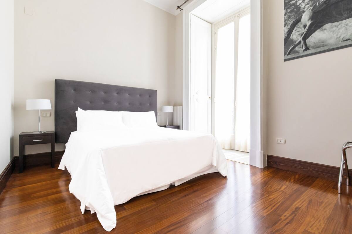 A bedroom with a large bed, two bedside tables with lamps, wooden flooring, and a large window with a view.