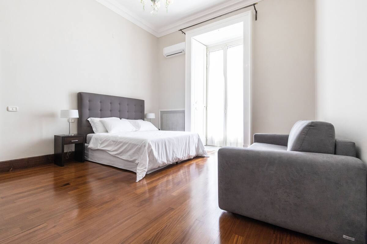 A modern bedroom with a bed, bedside tables, a grey sofa, and a large window.