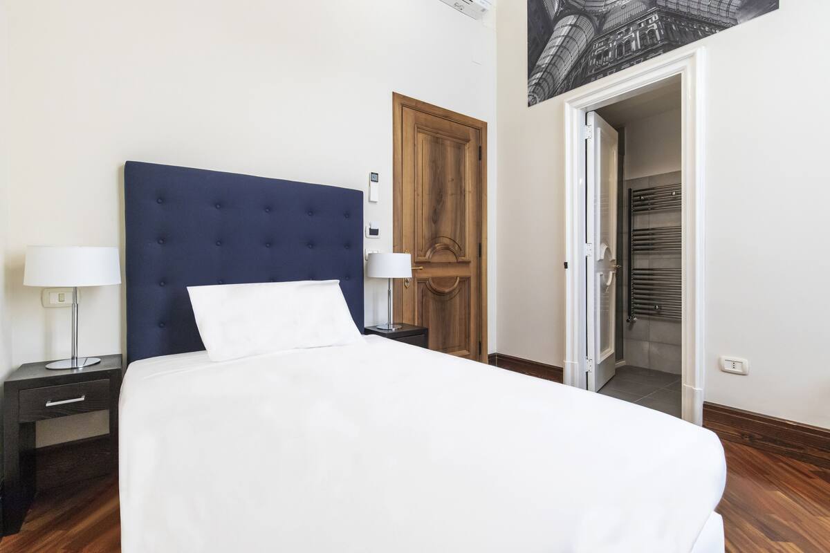 A neatly made bed with a blue headboard, two bedside tables with lamps, a wooden door, and a view of a bathroom with a towel rack.