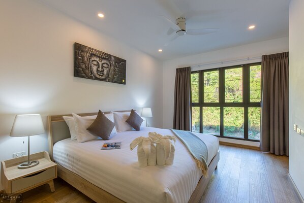 Une chambre à coucher avec un lit, des oreillers, une table de chevet, une lampe, un ventilateur de plafond et une fenêtre avec des rideaux.
