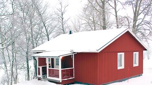 Une cabane en bois rouge, dotée d’un porche blanc et d’un toit recouvert de neige, entourée d’arbres.