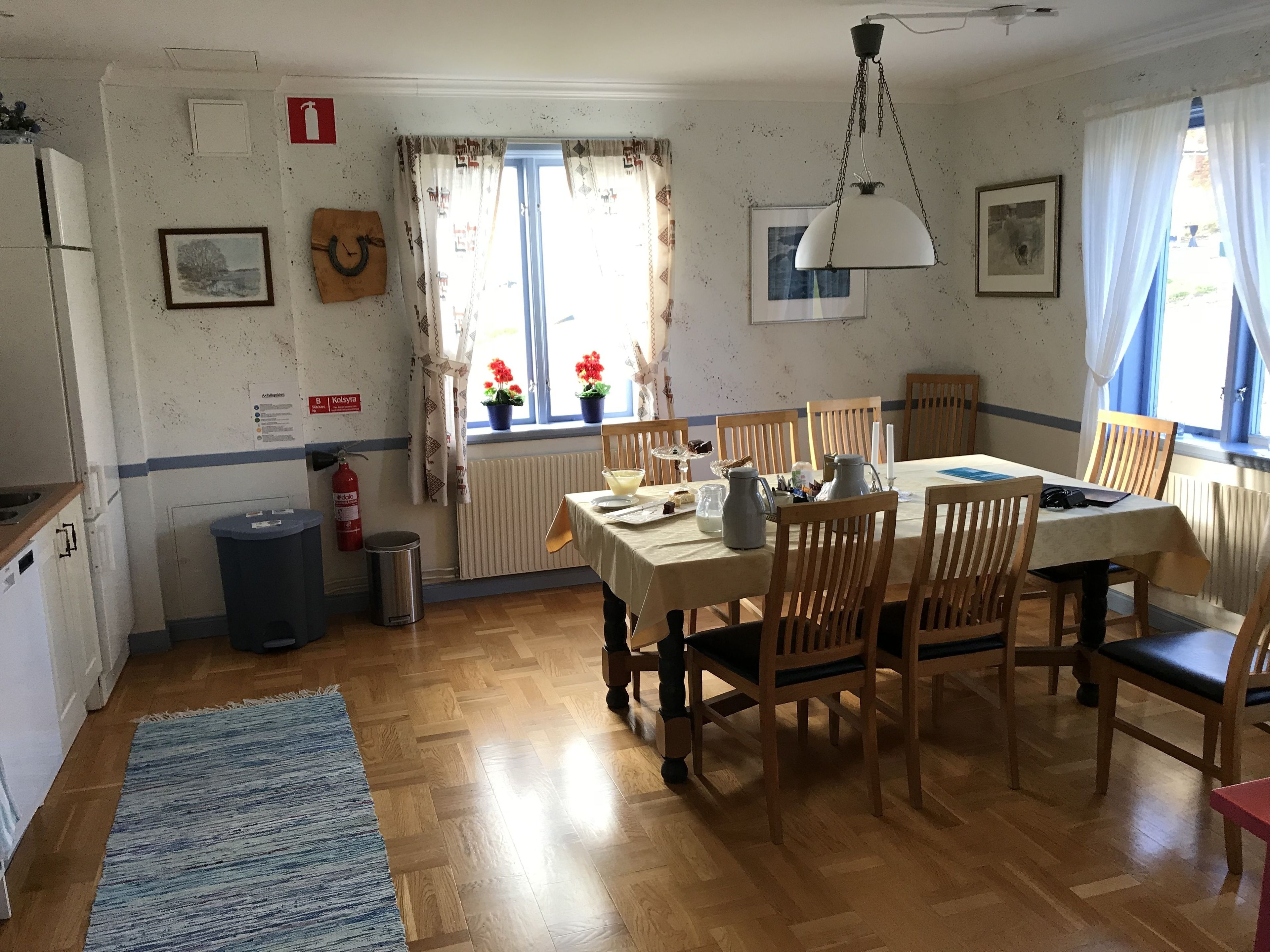 A dining area with wooden chairs and a table, a radiator, a fire extinguisher, and a window with curtains.