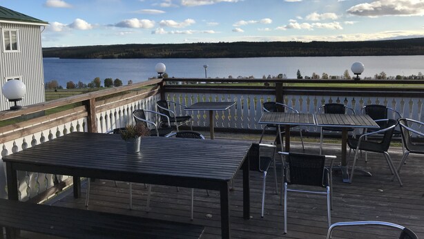 Une terrasse avec des sièges extérieurs donnant sur un lac, un bâtiment blanc avec un toit végétalisé et un ciel dégagé parsemé de nuages.