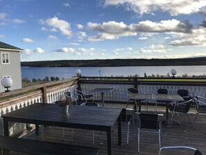A deck with outdoor seating overlooking a lake, a white building with a green roof, and a clear sky with clouds.