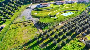 Vista aérea de una propiedad con varias construcciones, incluyendo una casa grande de color blanco con techo rojo, rodeada de campos verdes y olivares.