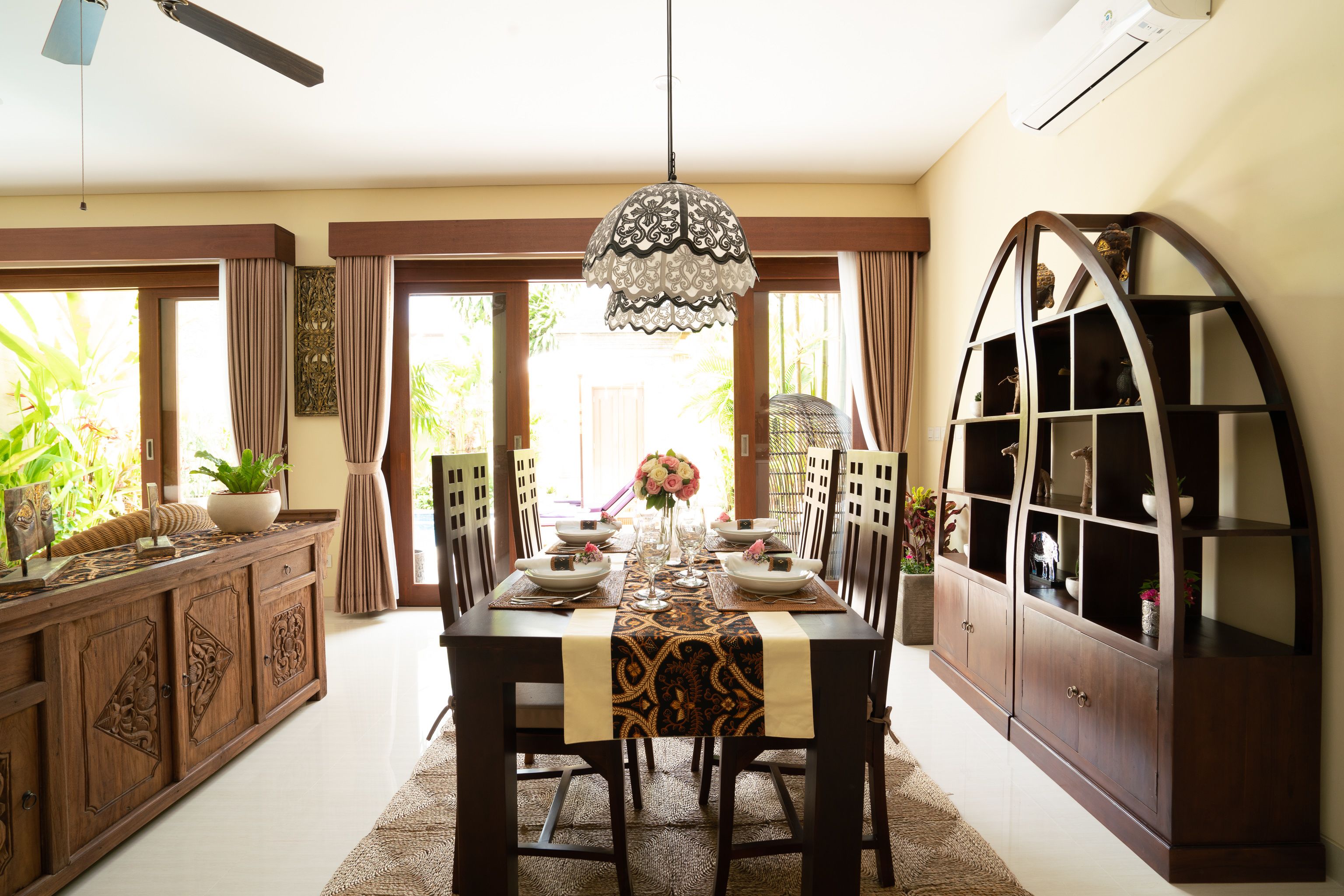 A dining area with a wooden table set for six, surrounded by chairs. There is a decorative hanging light fixture above the table.
