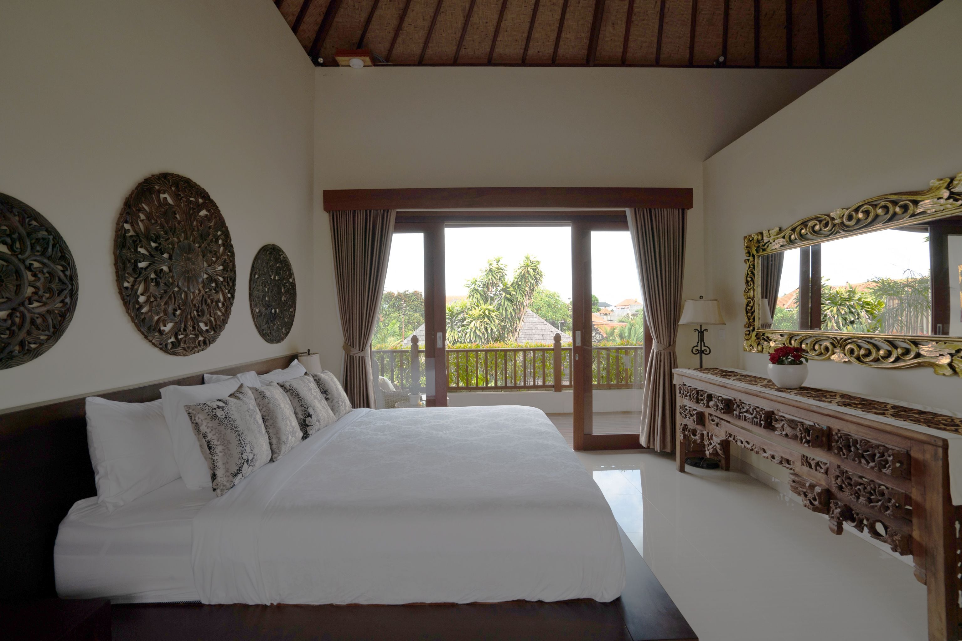 A bedroom with a large bed, a wooden console table, and a balcony with a view of palm trees.