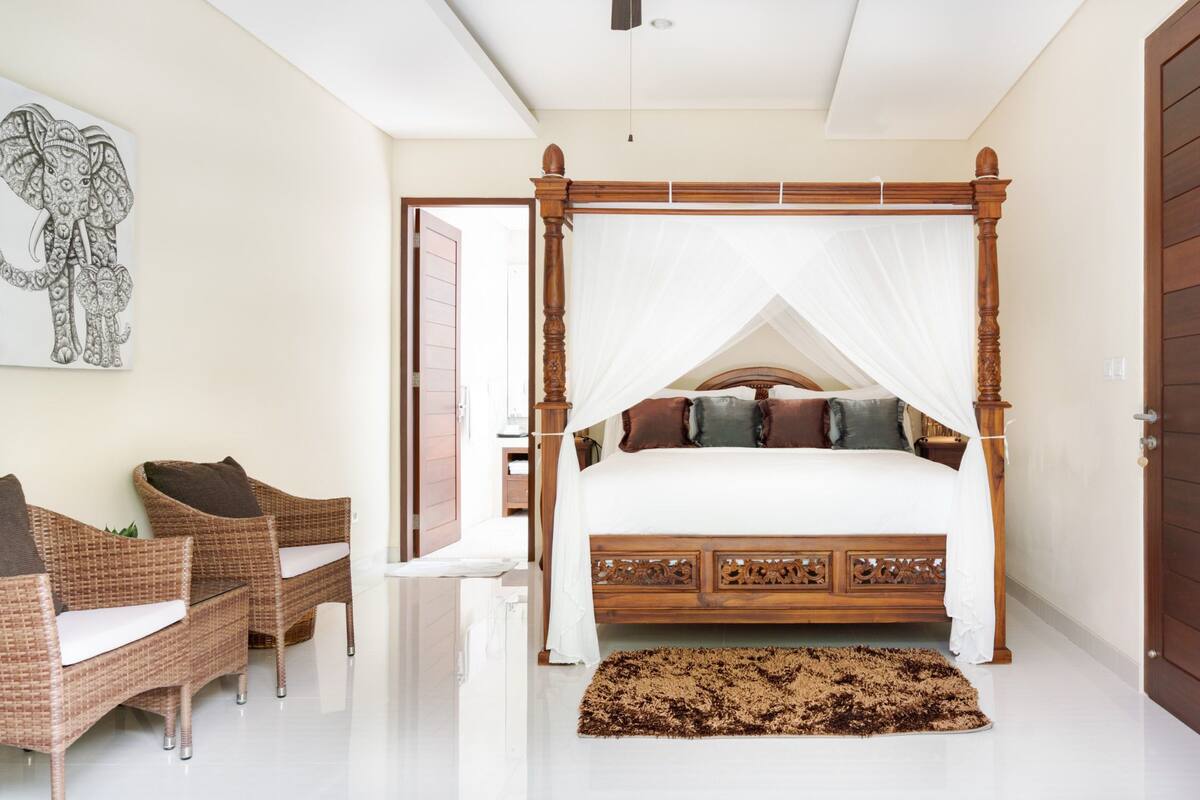A bedroom with a four-poster bed, wicker chairs, and a large elephant artwork on the wall.