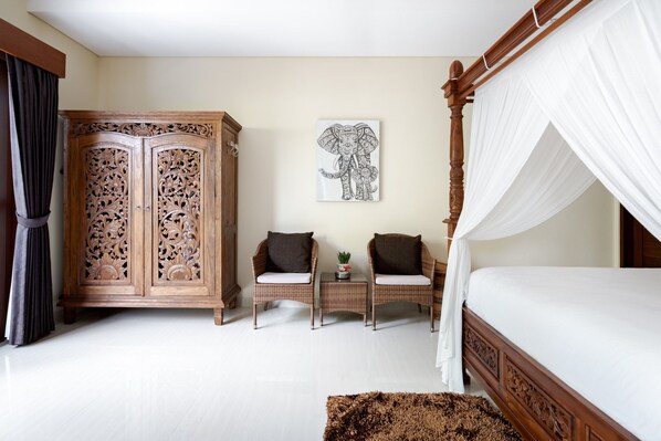 A bedroom with a four-poster bed, a wooden wardrobe, two wicker chairs, a small table with a plant, and an elephant artwork on the wall.