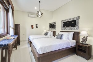 A hotel room with two beds, a wooden bench, decorative wall art, and a chandelier.