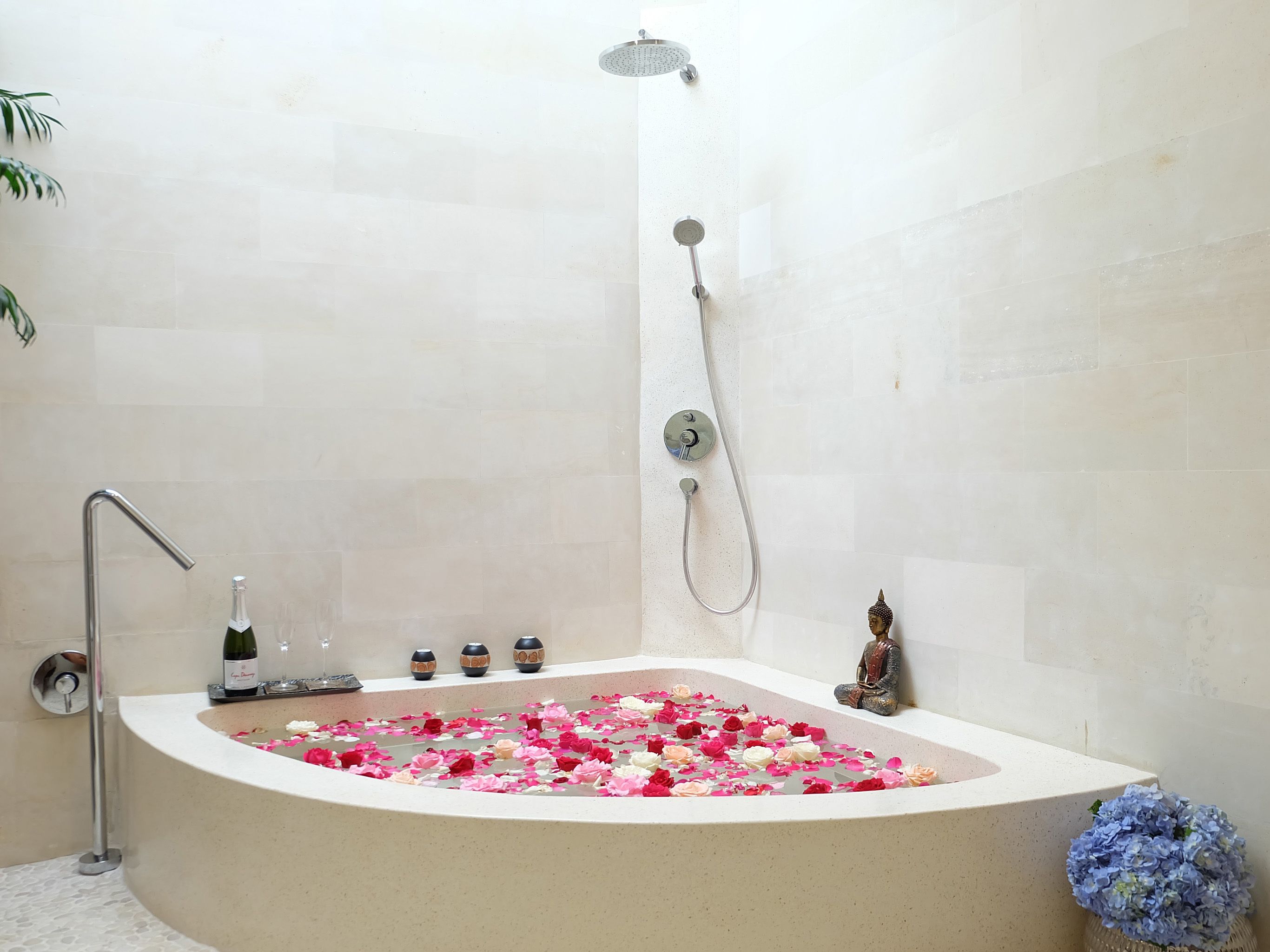 A modern bathroom with a freestanding bathtub filled with rose petals, a showerhead, and a small Buddha statue.