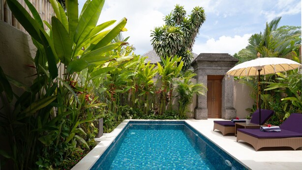 A rectangular swimming pool with a tiled blue surface, surrounded by lush tropical plants and a stone building with a door. - Kubu Gajah Villas (Denpasar)