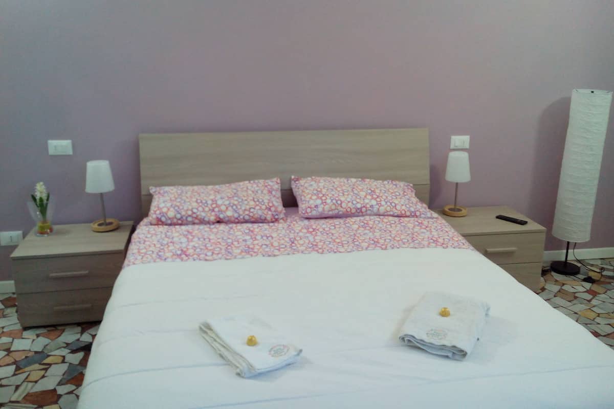 Hotellihuone, jossa on sänky, kaksi tyynyä, kaksi yöpöytää valaisimineen, yöpöydän päällä oleva maljakko, jossa on kukkia, ja suuri ikkuna, jossa on verho.