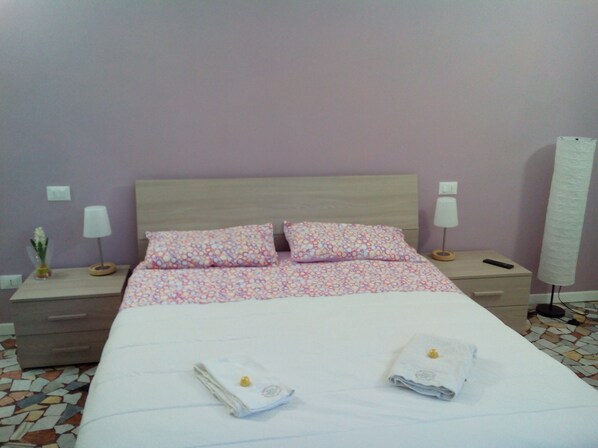 A hotel room with a bed, two pillows, two bedside tables with lamps, a nightstand, a vase with flowers, and a large window with a curtain.