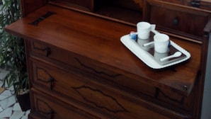 A wooden dresser with a tray holding two cups and a spoon, a remote control on top, and a wicker wall hanging. - Il Cantuccio B&B (Bologna)