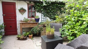 Een klein, omheind tuinperk met een rode deur, rieten meubels en verschillende planten in potten.