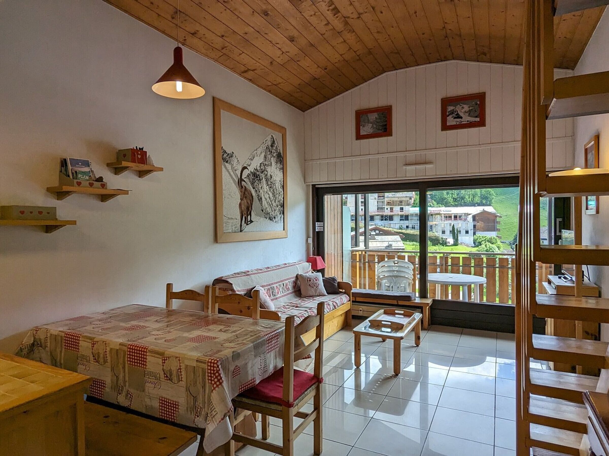 Una habitación acogedora con techo de madera, un ventanal con vistas a un paisaje verde, un sofá, una mesa y una lámpara colgante.