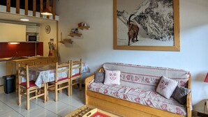 Una sala de estar acogedora con techo de madera, un comedor con mesa y sillas de madera, y un sofá con cojines estampados.
