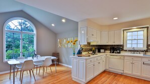A modern kitchen with white cabinetry, a dining area with a table and chairs, and a large window with a view of greenery.