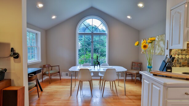 A dining area with a large window, wooden flooring, white chairs, and a white table.