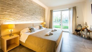 A hotel room with a large bed, a nightstand, a lamp, a window with curtains, and a view of greenery outside. - Agriturismo Dolceluna (Milan)