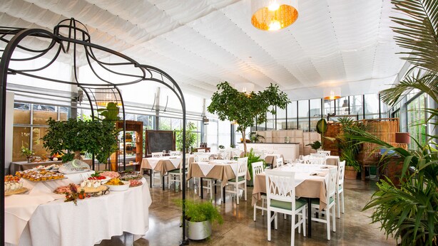 A spacious indoor dining area with tables set for a meal, surrounded by potted plants and natural light from large windows. - Agriturismo Dolceluna (Milan)